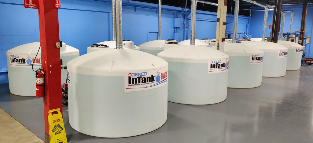 A row of large, white industrial tanks labeled "SCIENO InTank" in a clean, blue-walled facility. A fire extinguisher and caution sign are visible.