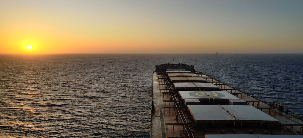 Sunset over the ocean with a large cargo ship in the foreground. The calm sea and warm hues of the sky create a tranquil, serene atmosphere.