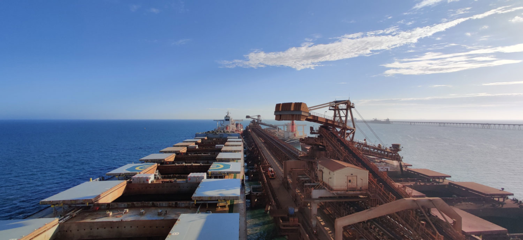 Large cargo ship with open holds alongside an industrial loading dock, set against a vast blue ocean and clear sky.