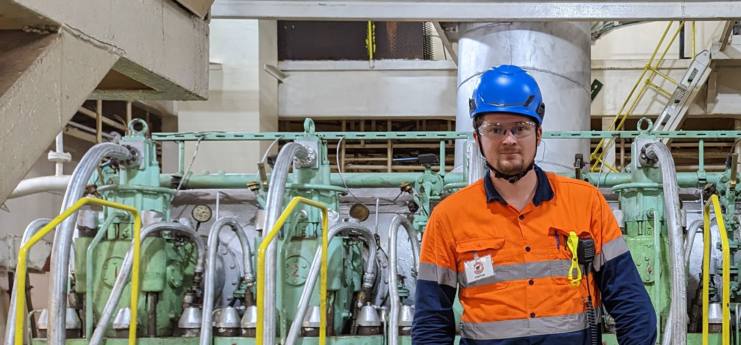 A worker in a bright orange vest and blue hard hat looking at the camera and smiling, while standing in front of green pipes and machinery.