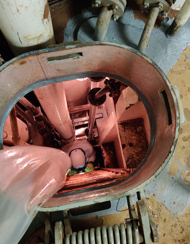 View looking down through a circular hatch into a confined industrial space where a worker in protective gear and a hard hat is positioned among pipes and mechanical components.